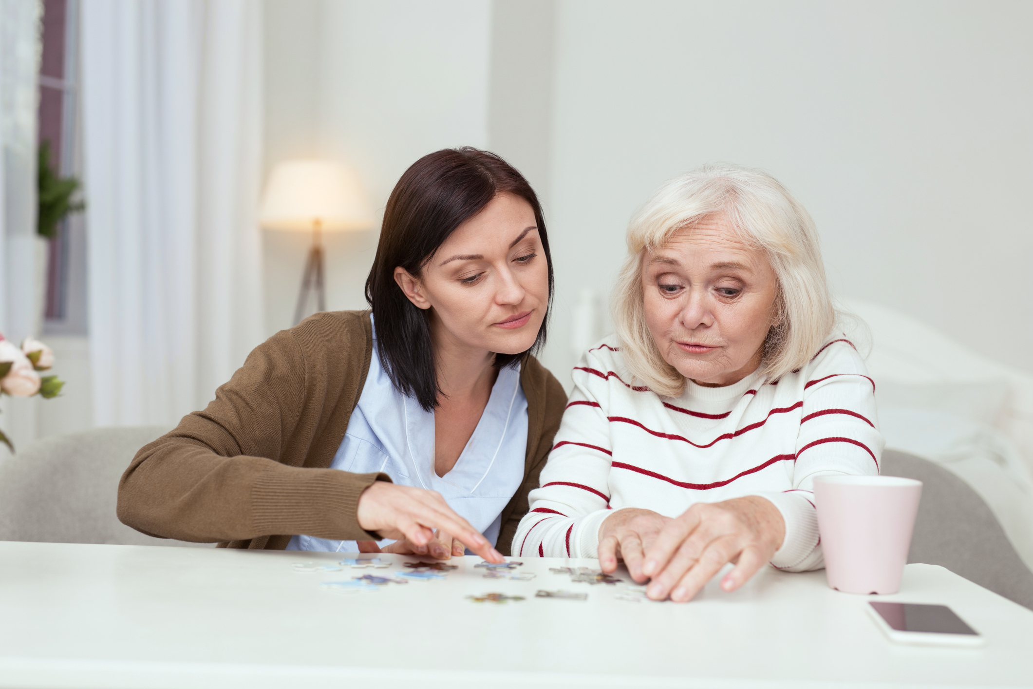 Pleasant time spending. Jolly elder woman and caregiver sitting while gathering puzzle